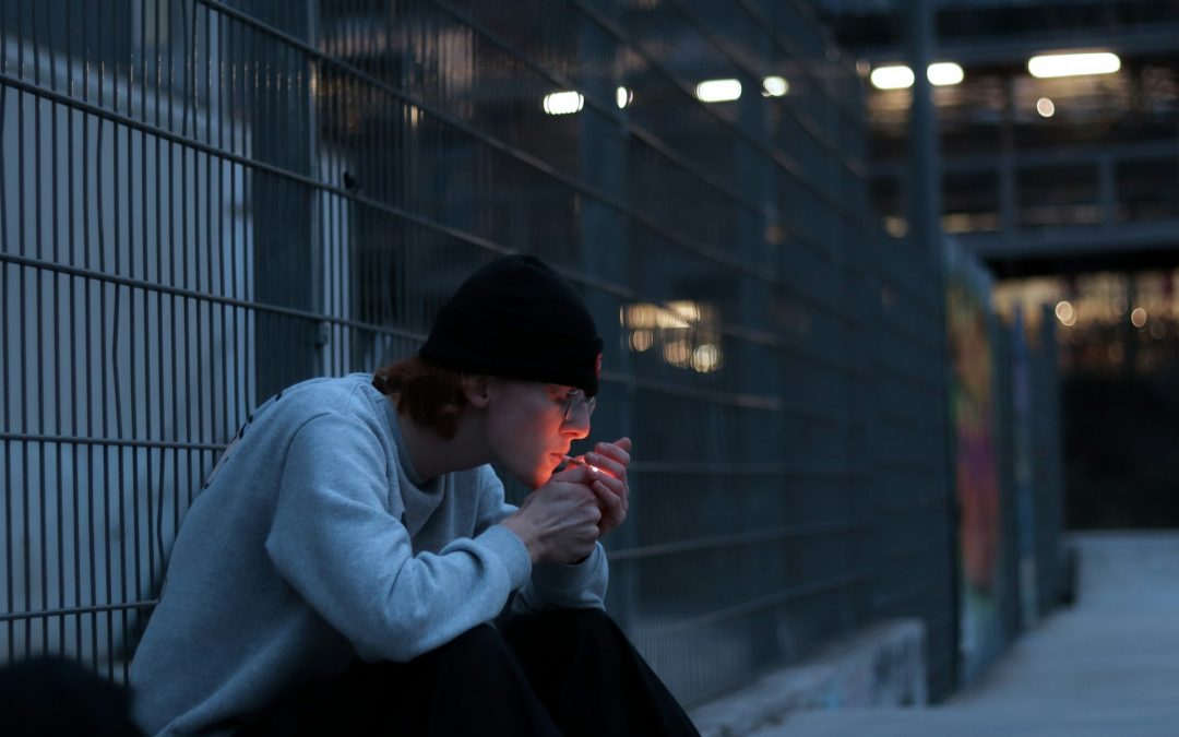 This image captures a young person sitting on the ground next to a metal fence, lighting a cigarette in the dim light of early evening. The person is dressed in a casual gray sweatshirt and black beanie, absorbed in the act. The background features a blurred urban setting with soft lighting and a mural on a distant wall, contributing to a somber mood.