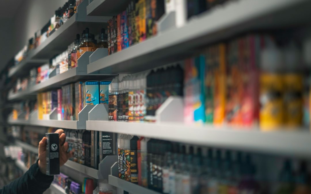 Close-up of a person's hand selecting a vaping product from a blurred background displaying a wide range of colorful e-liquid bottles on retail shelves (Photo by E-Liquids UK on Unsplash)
