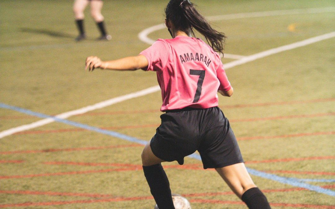 A girl in a pink football jersey and black shorts is about to kick a ball on an indoor football field. Her jersey has the name "Amaarah" and the number 7 on the back. Other players are visible in the background.