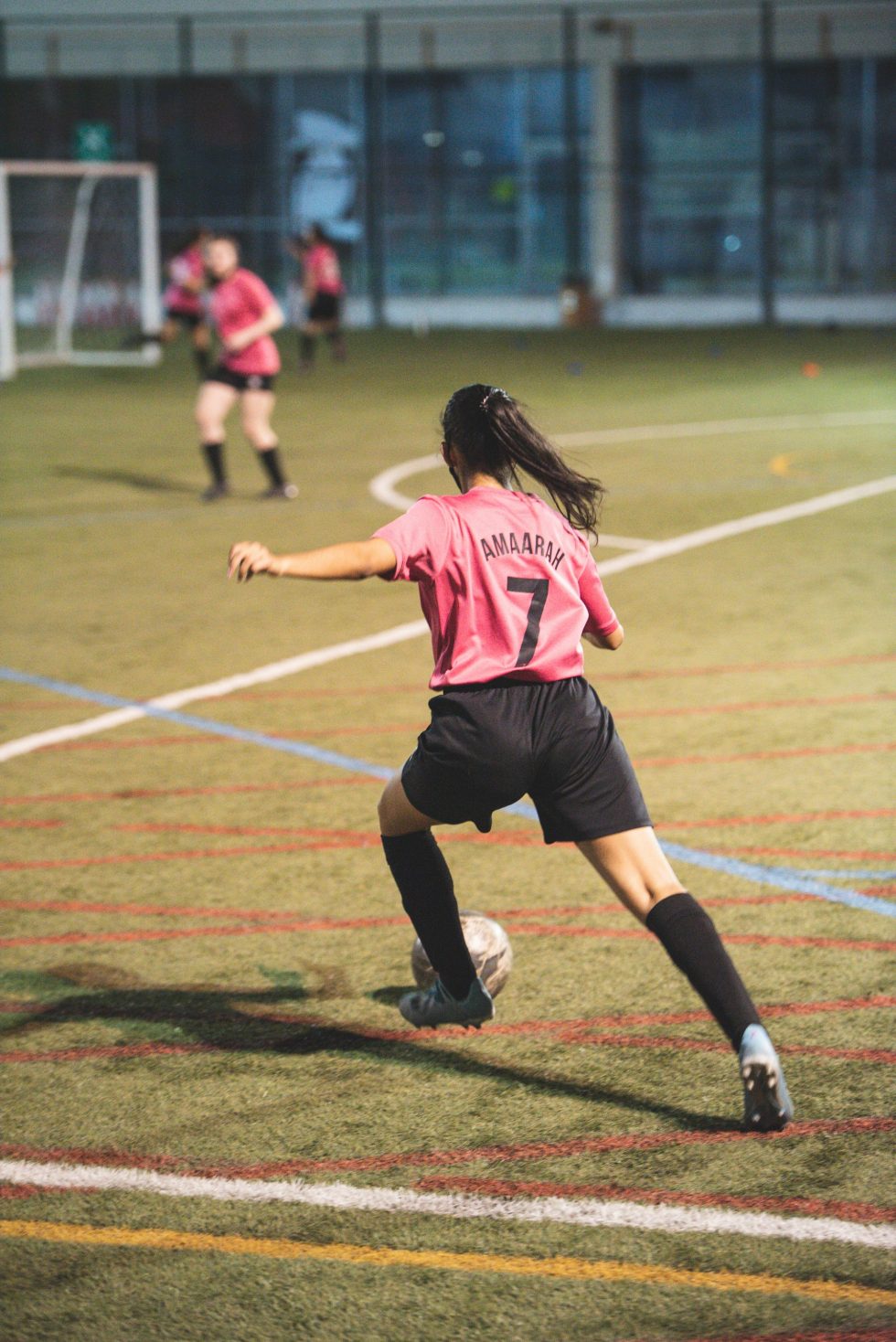 A girl in a pink football jersey and black shorts is about to kick a ball on an indoor football field. Her jersey has the name 