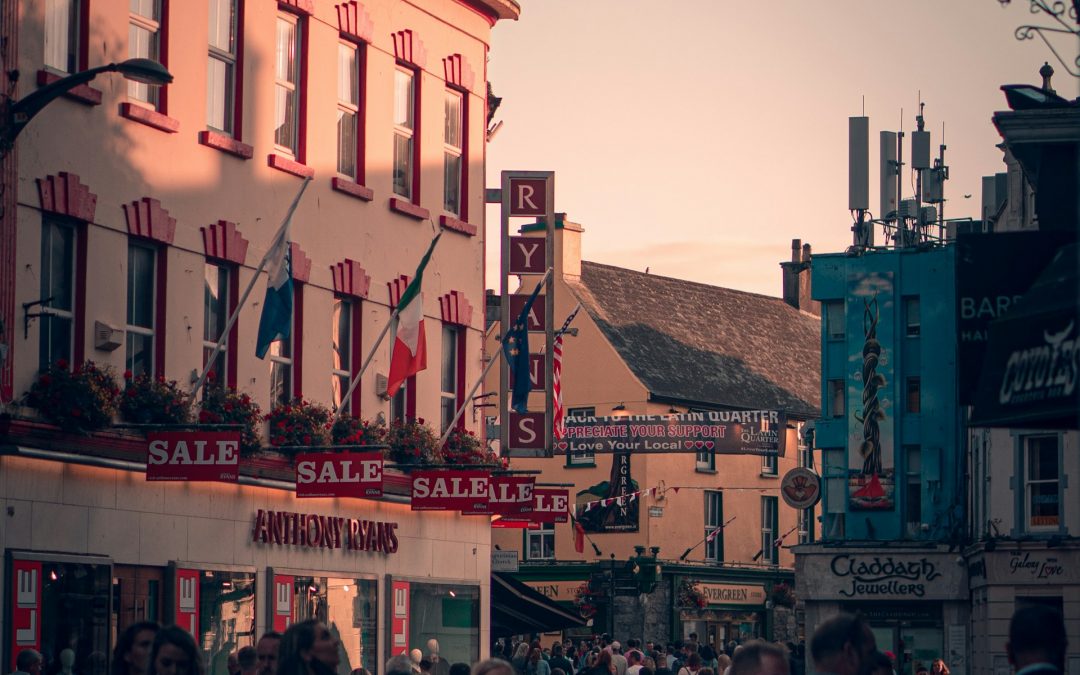 This image depicts a bustling street scene in Galway, Ireland during the evening. The warm golden light of sunset illuminates the façades of traditional buildings adorned with multiple signs, including ones for sales and local businesses. People of various ages are seen walking, shopping, and socialising.
