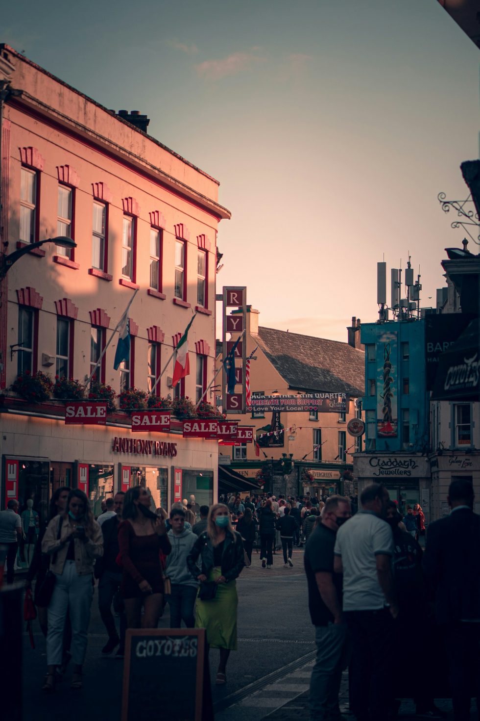 This image depicts a bustling street scene in Galway, Ireland during the evening. The warm golden light of sunset illuminates the façades of traditional buildings adorned with multiple signs, including ones for sales and local businesses. People of various ages are seen walking, shopping, and socialising.