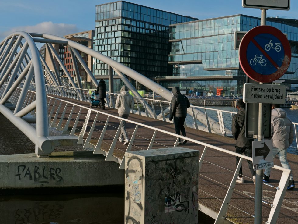 Young Dutch people walking on a modern bridge with a 'no cycling' sign and urban buildings in the background.