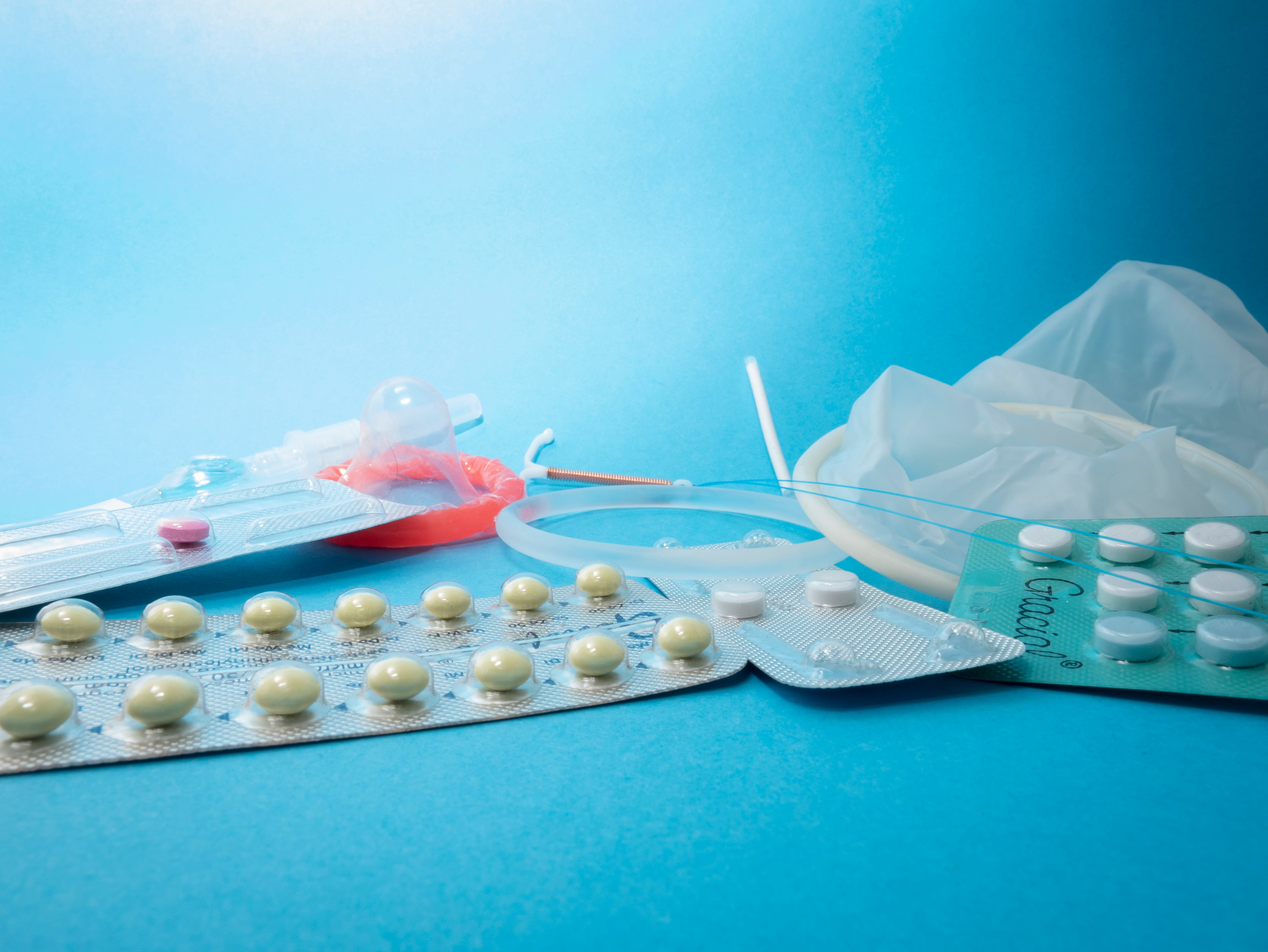 A selection of contraceptives displayed on a blue background, including contraceptive pills in blister packs, a condom in its packaging, a contraceptive implant, an intrauterine device (IUD), and a diaphragm.