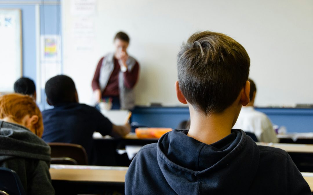 Rear view of a classroom with students seated at desks, focusing on a teacher standing in front of a whiteboard in the background.